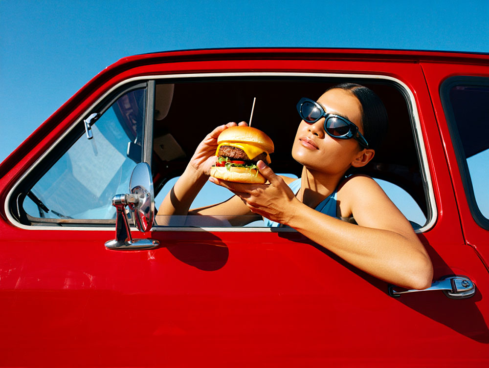 Jeune femme lunettes noires dévorant un burger dans décapotable rouge, rendu photoréaliste par Reve Image 1.0.