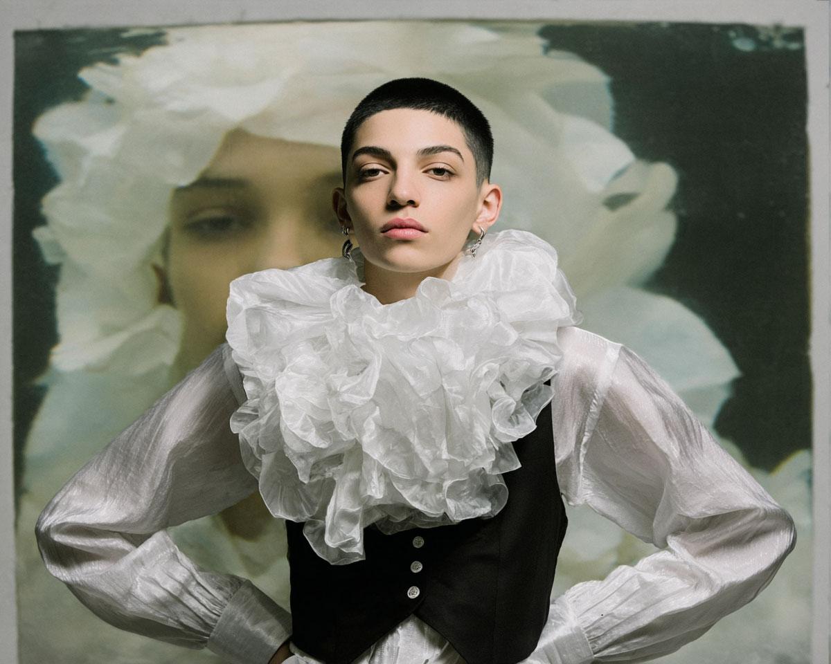 Portrait d’un créatif androgyne en chemise blanche à col plissé, son visage projeté en arrière-plan, représentant le dialogue intérieur entre humain et image.