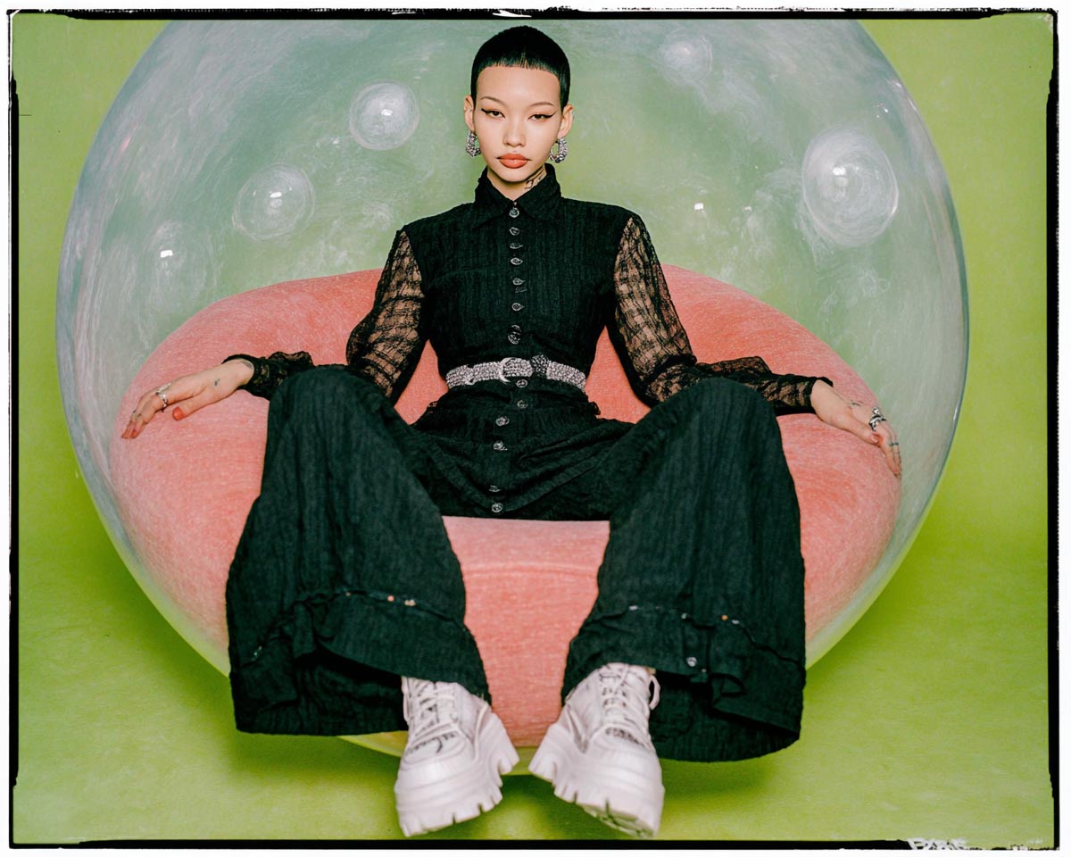 Créatrice vêtue de noir assise dans une bulle transparente sur un fauteuil rose, posée dans un décor vert clair, illustrant le calme et l’introspection créative à l’ère de la retouche photo IA.
