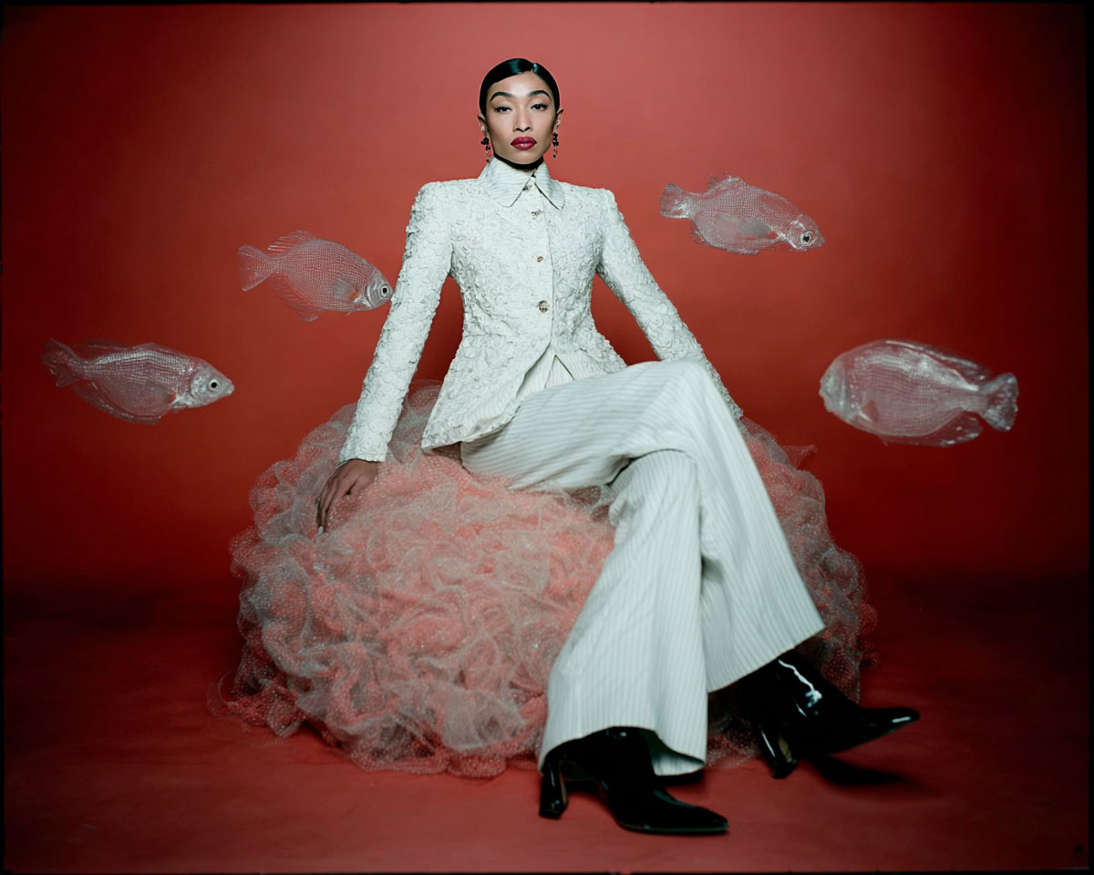 Portrait d’une créatrice en costume clair assise sur une structure rose, entourée de poissons translucides flottants dans un espace rouge, symbolisant le futur de la création visuelle avec IA.